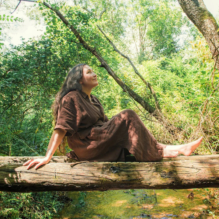 A person enjoying nature while sitting on a log in a forested area, surrounded by greenery and sunlight.